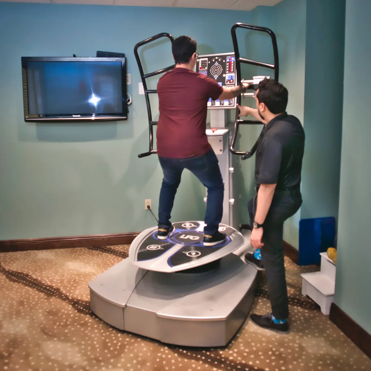 A man standing on the Spine Force platform holding the support handles while a clinician assists at the console